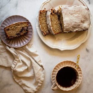 A slice of gluten free cherry loaf cake on a plate, the loaf next to it, and a cup of tea.