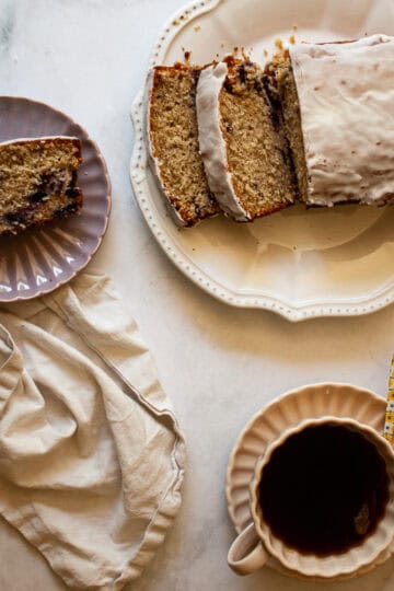 A slice of gluten free cherry loaf cake on a plate, the loaf next to it, and a cup of tea.