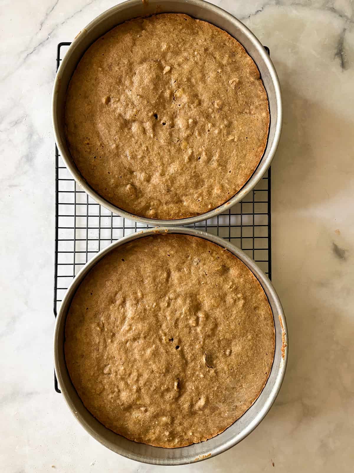 This gluten-free hummingbird cake is a moist, fruit-infused cake topped with tangy cream cheese icing and sprinkled with toasted walnuts. A gluten free hummingbird cake cools in the pans.