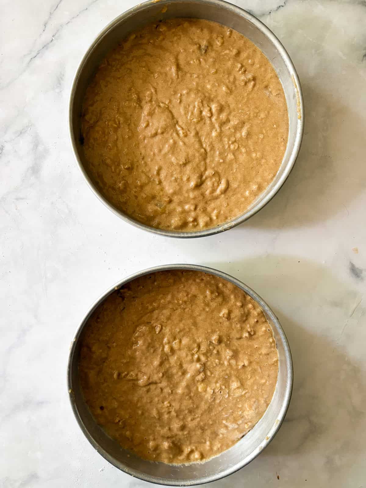 This gluten-free hummingbird cake is a moist, fruit-infused cake topped with tangy cream cheese icing and sprinkled with toasted walnuts. Gluten free hummingbird cake batter rests in 2 cake pans.