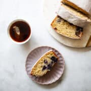 A cup of tea, a slice of lemon blueberry loaf on a plate, and the sliced loaf.
