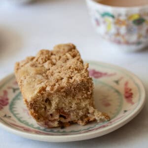 A piece of gluten free rhubarb crumb cake on a plate with a cup of tea behind it.