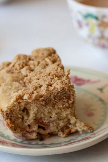 A piece of gluten free rhubarb crumb cake on a plate with a cup of tea behind it.