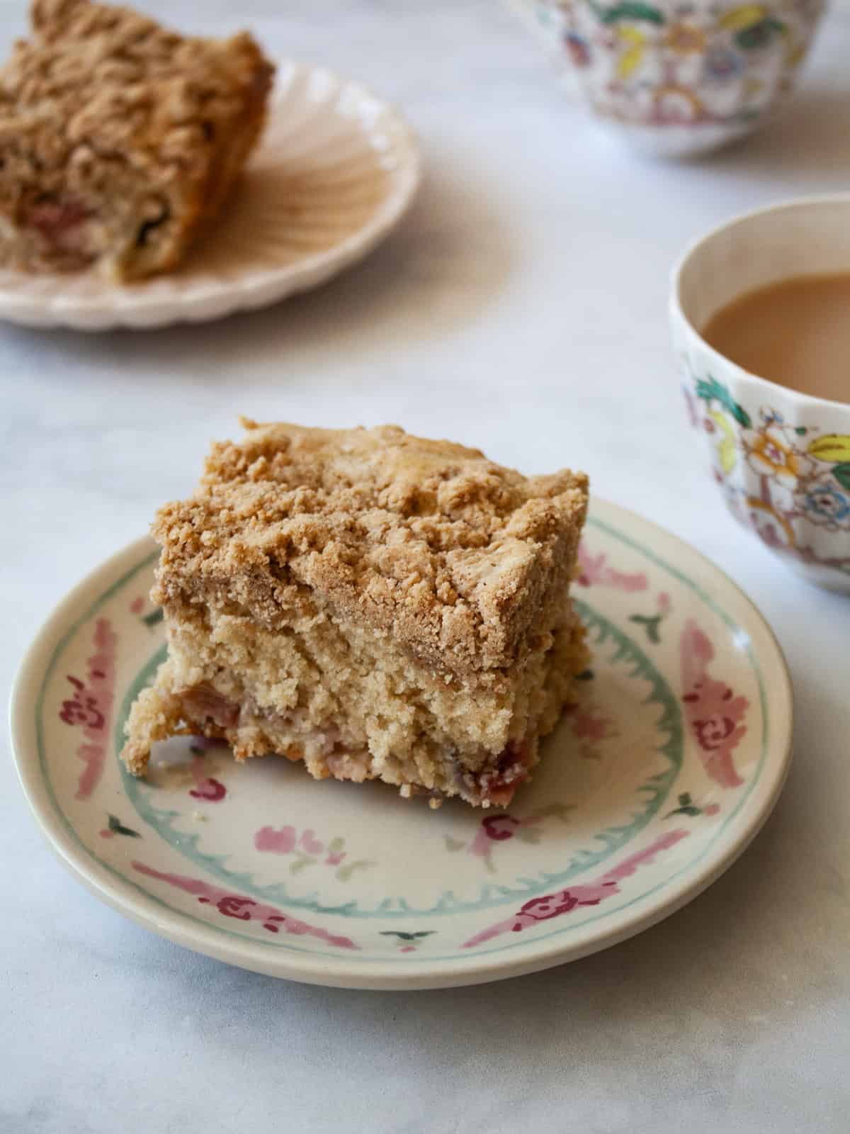 Pieces of gluten free rhubarb crumb cake on a plate with a cup of tea behind it.