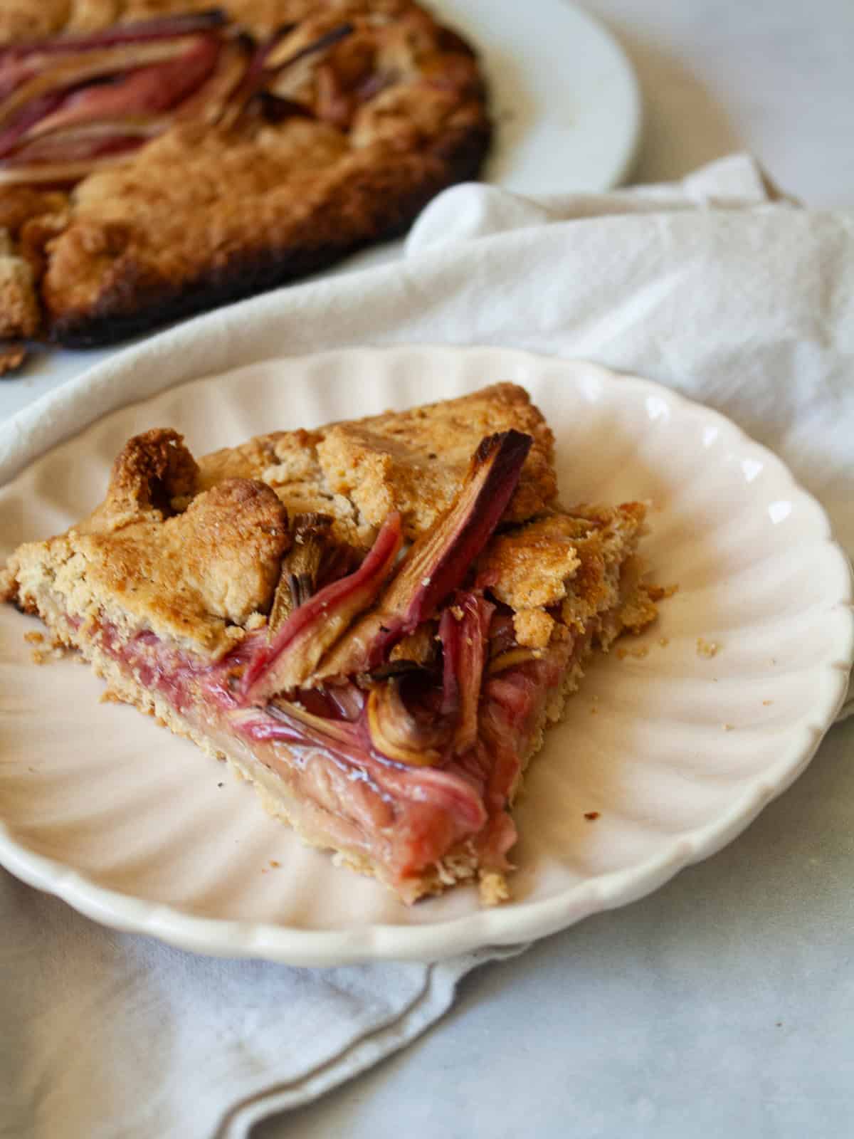 A tender and flaky gluten-free pastry wraps around sweet-tart slices of rhubarb in this rustic gluten free rhubarb galette. A slice of freshly baked gluten free rhubarb galette on a plate.