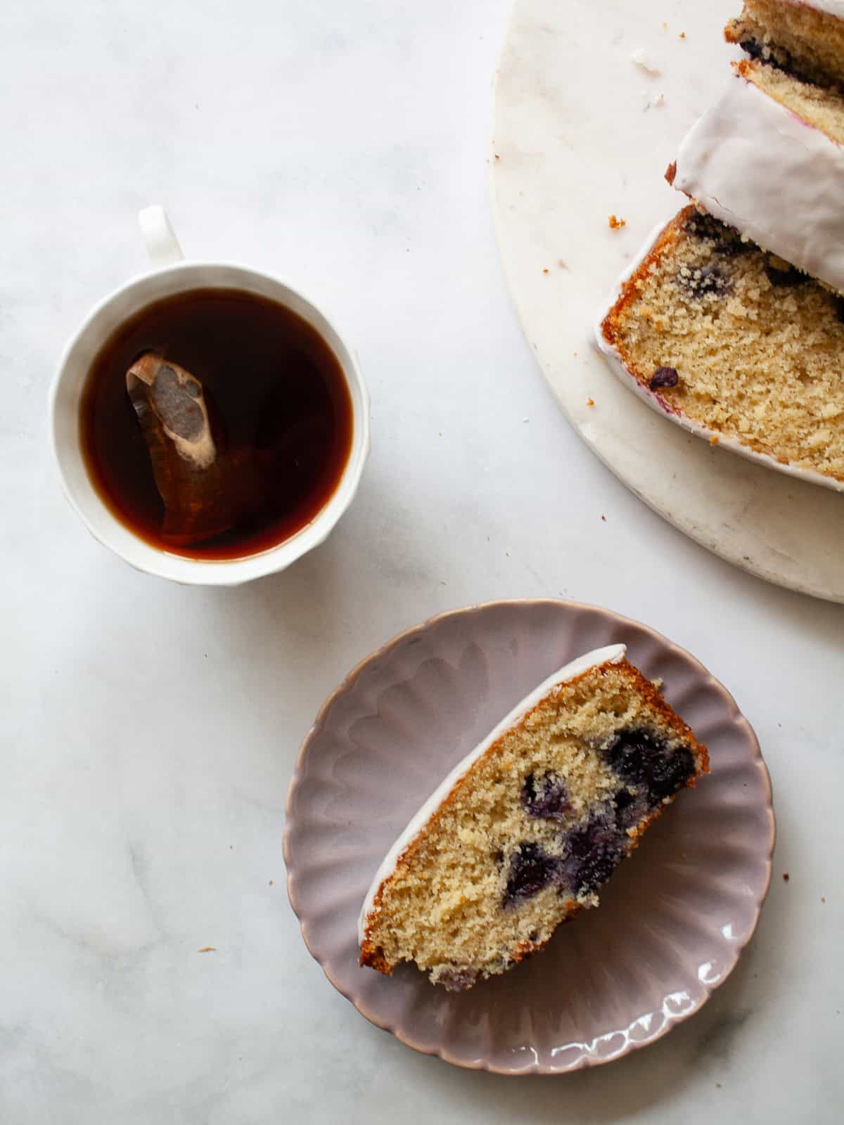 A soft and sweet lemon blueberry loaf that's studded with juicy fruit and topped with a lusciously thick, lemon-spiked glaze. A cup of tea, a slice of lemon blueberry loaf, and the bread nearby.