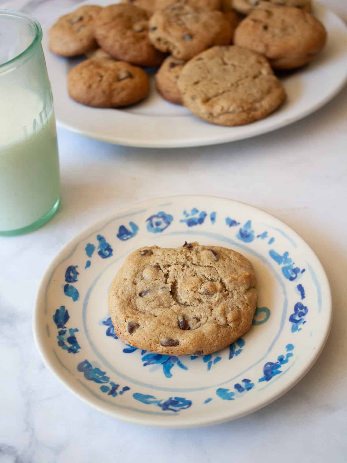 One of my favorite types of cookies, these maple chocolate chip cookies are sweetened with a combination of maple syrup and brown sugar. A glass of milk next to a plate with a maple syrup chocolate chip cookie.