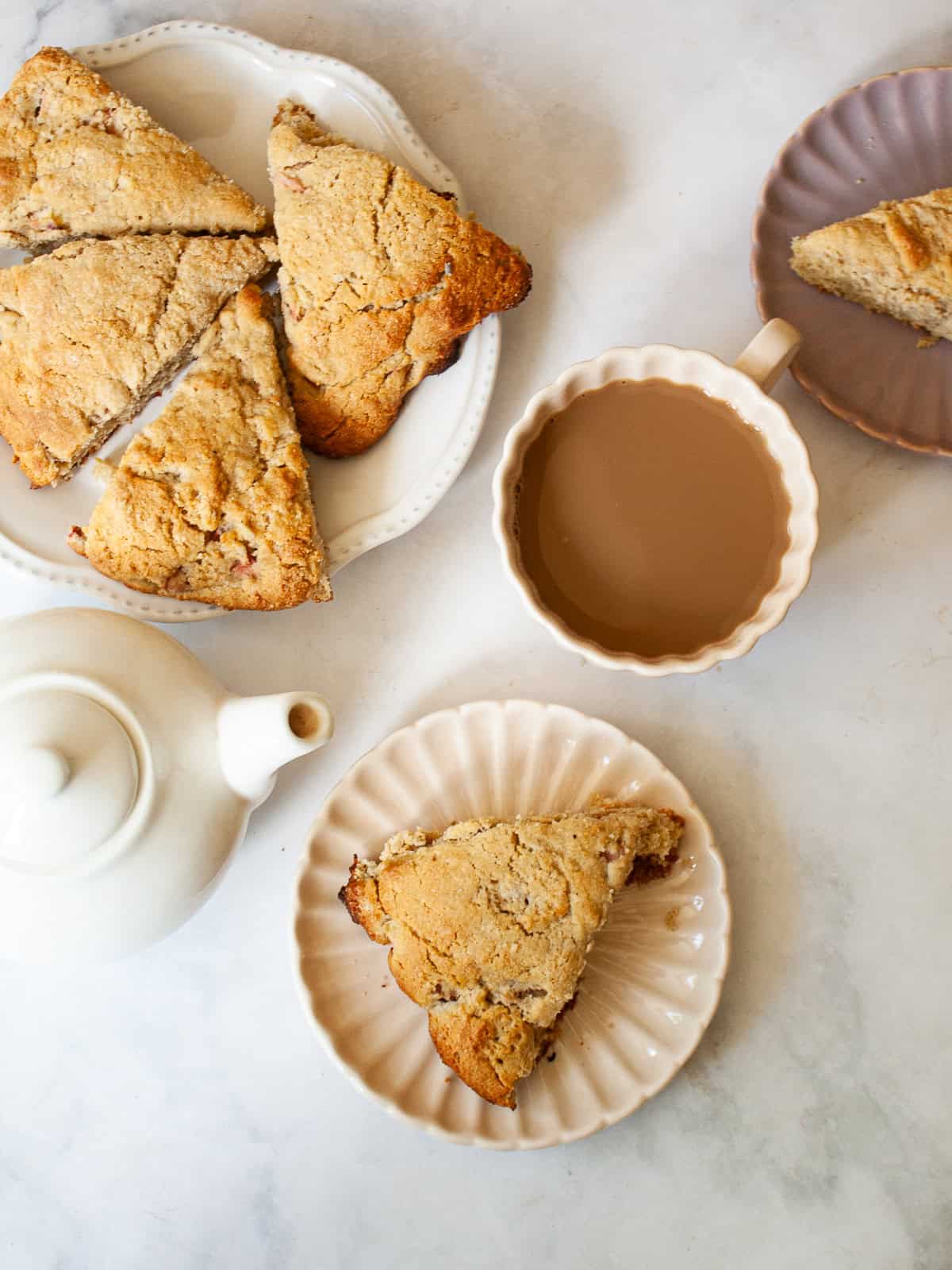 Whole-grain oat flour rhubarb scones are naturally gluten-free, warmed with a bit of orange zest and studded with jammy pockets of rhubarb. A plate of oat flour rhubarb scones next to a pot of tea.