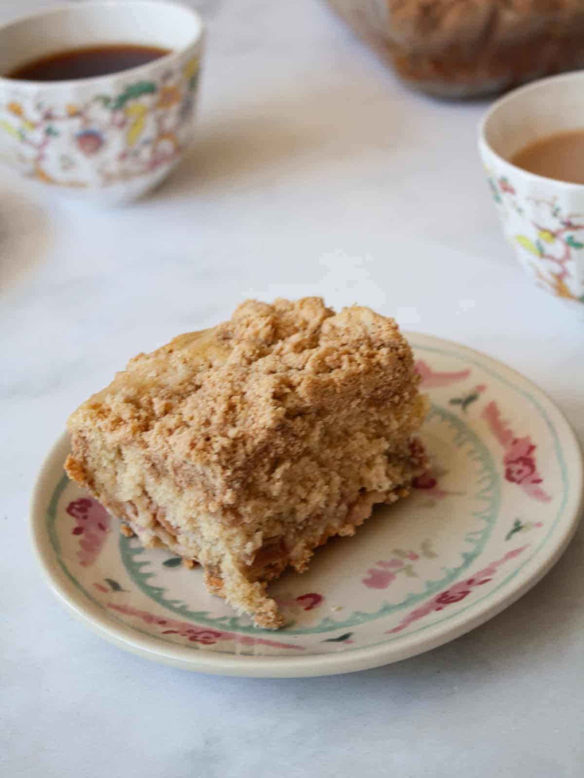 This rhubarb crumb cake is studded with jammy bits of tart rhubarb and topped with a cinnamon-laced brown sugar streusel. A portion of rhubarb crumb cake on a plate with cups of tea.