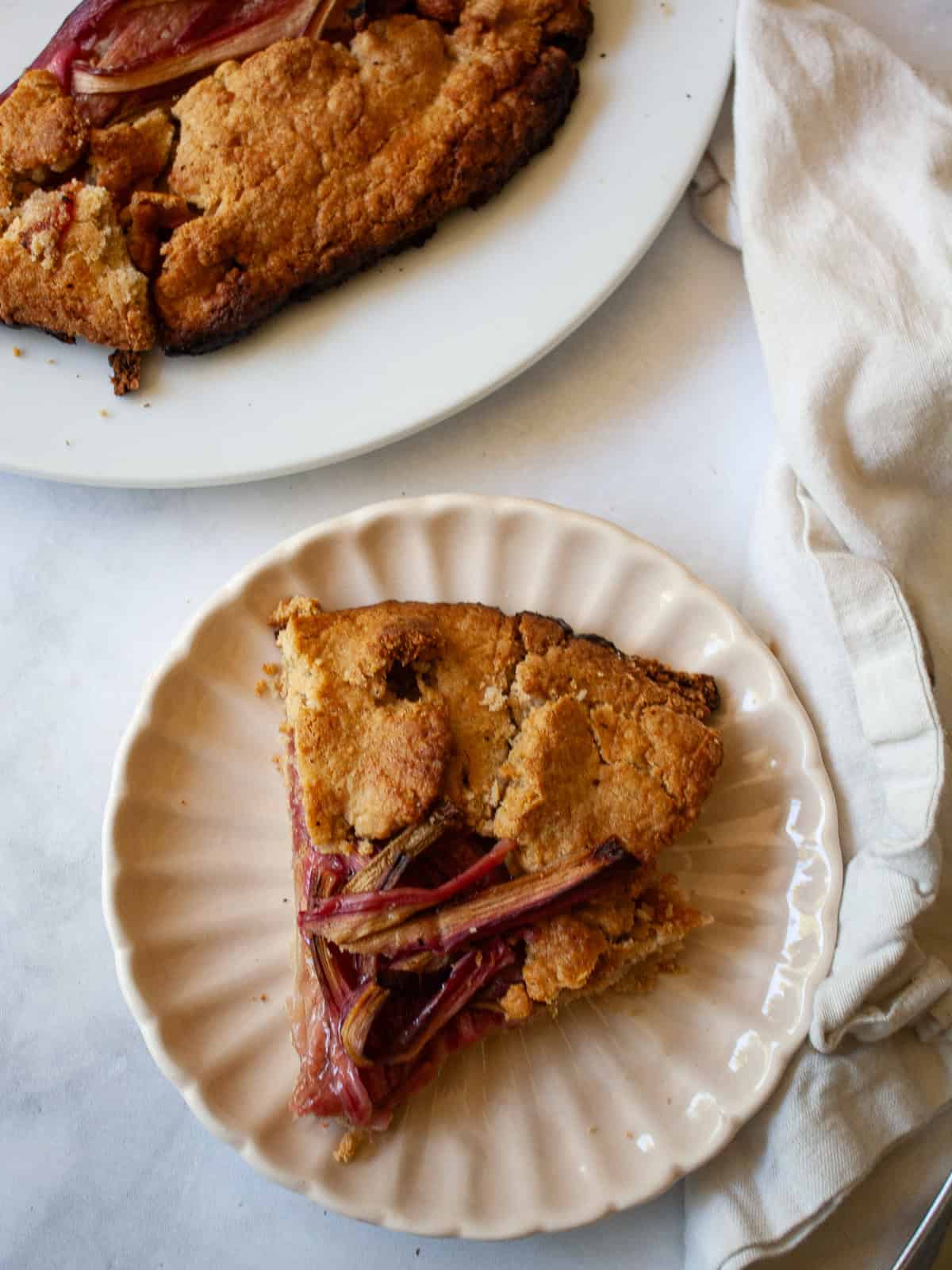 This rhubarb galette features a flaky, whole-grain crust folded around a jammy filling. Rhubarb's flavor truly shines in this rustic bake. A piece of rhubarb galette on a plate with the galette behind it.