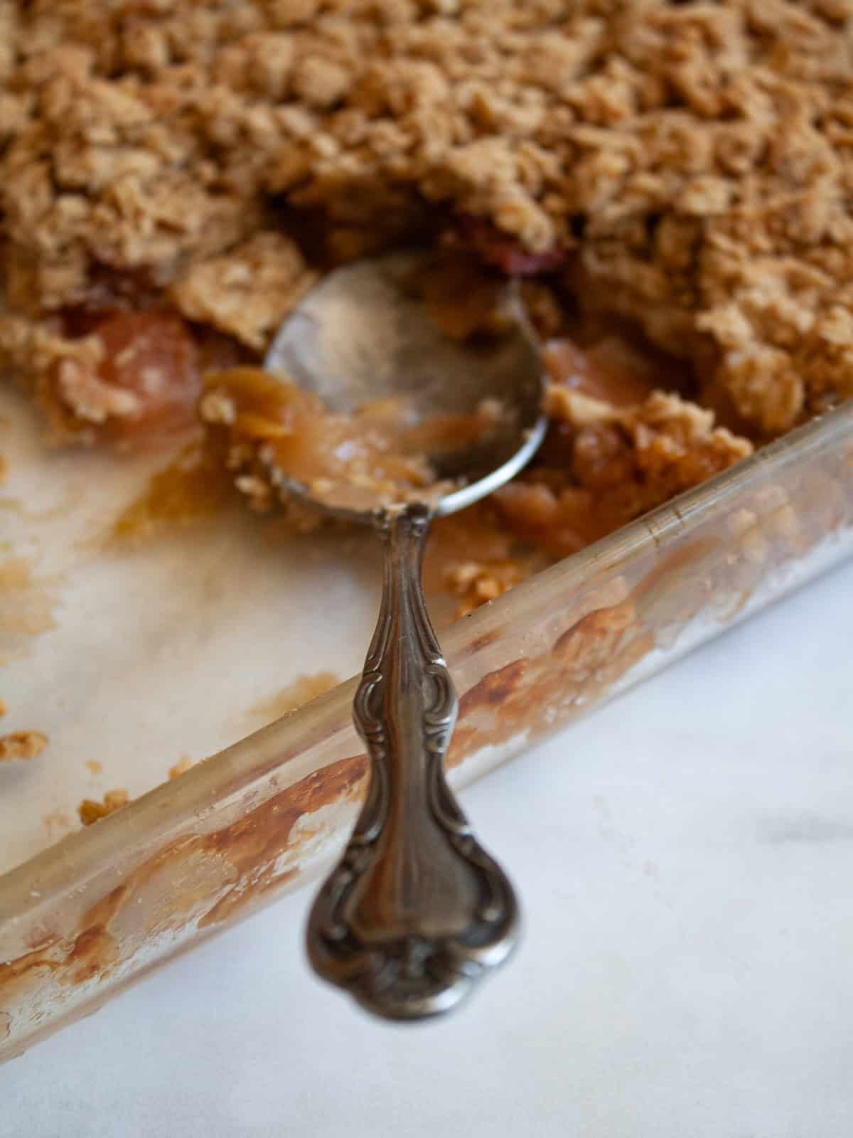 A jammy apple rhubarb crisp pairs seasonal rhubarb with sweet apples. Make the buttery oat topping gluten-free or use whole wheat flour. A serving spoon rests in a pan of rhubarb apple crisp.