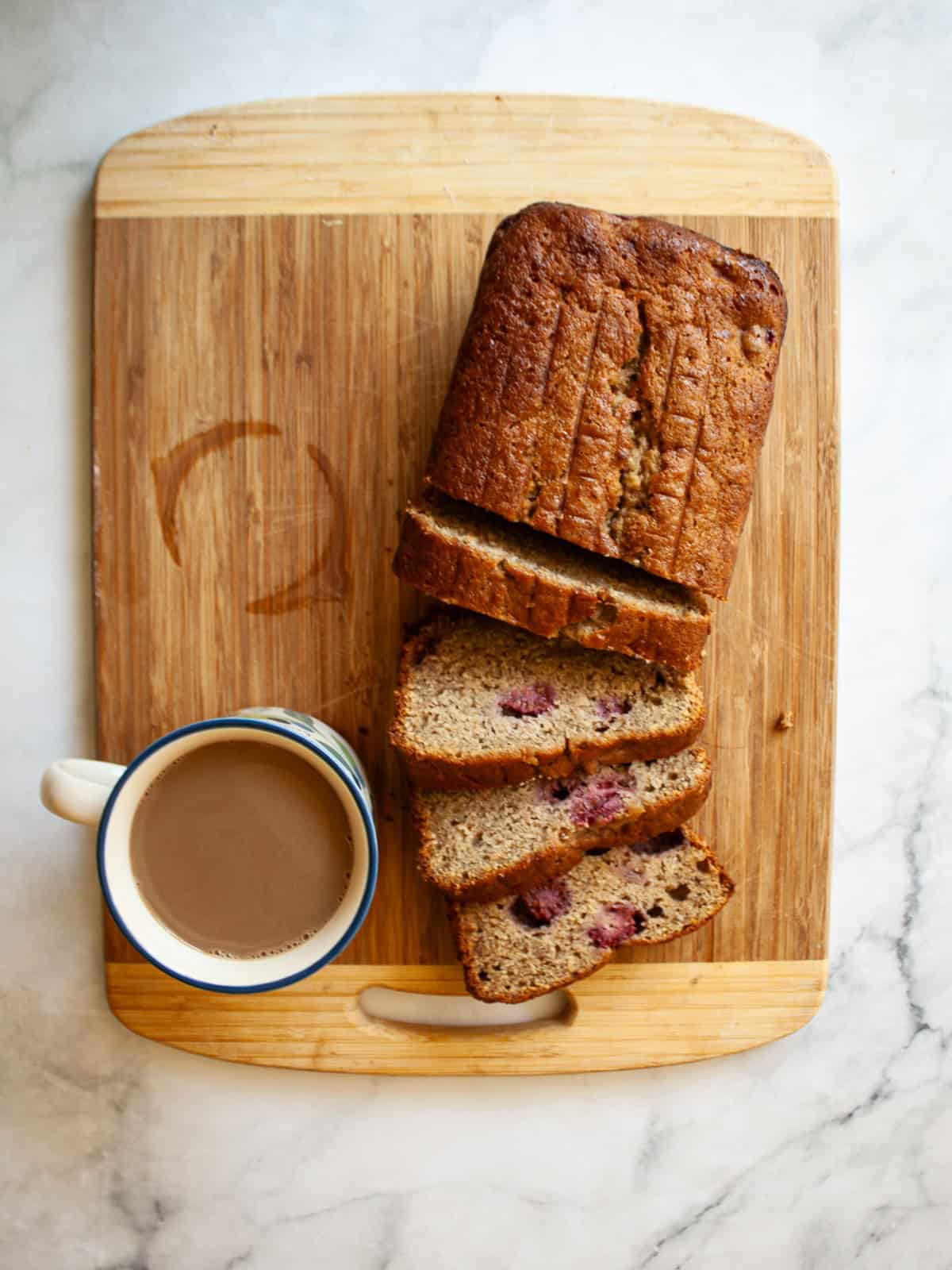 An uptick on a classic, this whole-grain raspberry banana bread features a soft, tender bread studded with juicy pockets of raspberries. A sliced loaf of banana raspberry bread on a cutting board .with a cup of coffee
