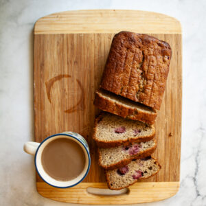 A sliced loaf of banana raspberry bread on a cutting board .with a cup of coffee