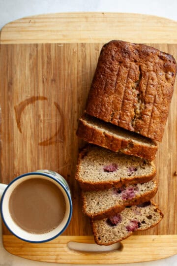A sliced loaf of banana raspberry bread on a cutting board .with a cup of coffee