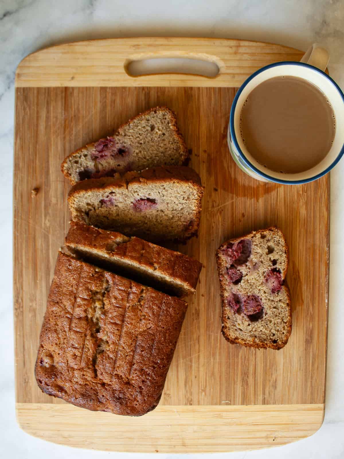 A cutting board with a sliced loaf of banana raspberry bread and a cup of coffee.