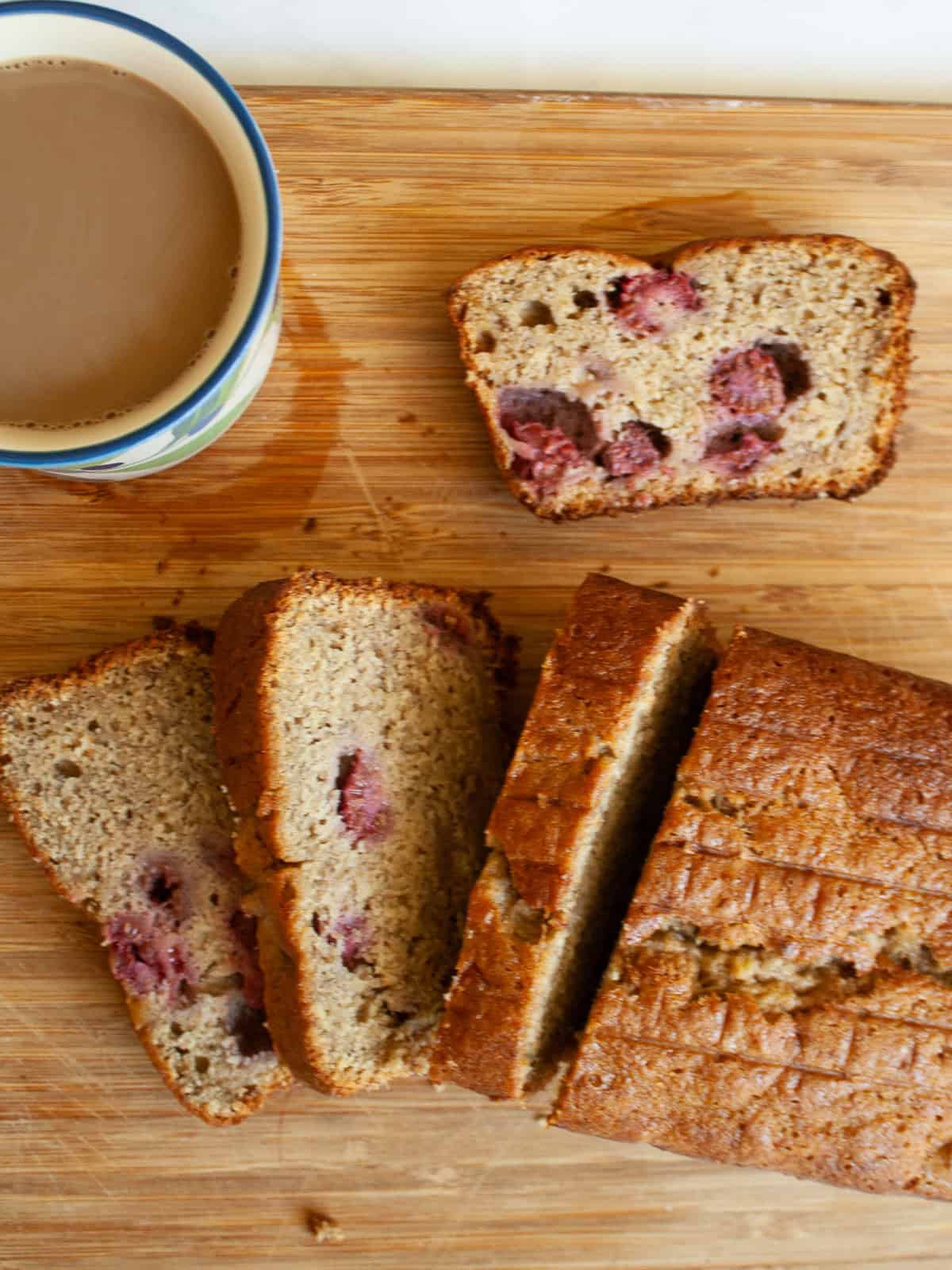 An uptick on a classic, this whole-grain raspberry banana bread features a soft, tender bread studded with juicy pockets of raspberries. A cup of coffee next to slices of banana raspberry bread.