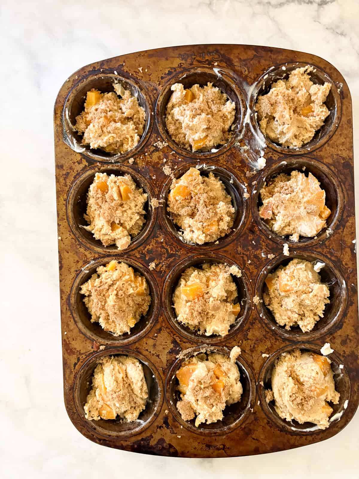Yogurt, whole grains, and juicy peaches are swirled together with brown sugar in these tender and good-for-you gluten free peach muffins. Gluten free peach muffin batter in a muffin tin.