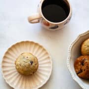 A gluten free raspberry muffin on a plate, a cup of coffee, and a bowl of muffins.