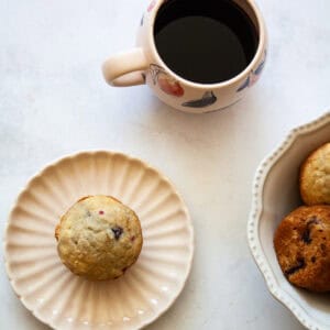 A gluten free raspberry muffin on a plate, a cup of coffee, and a bowl of muffins.