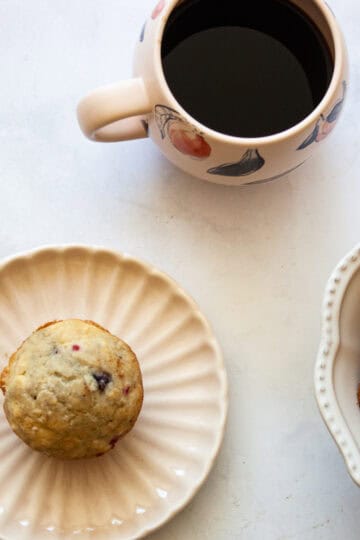 A gluten free raspberry muffin on a plate, a cup of coffee, and a bowl of muffins.