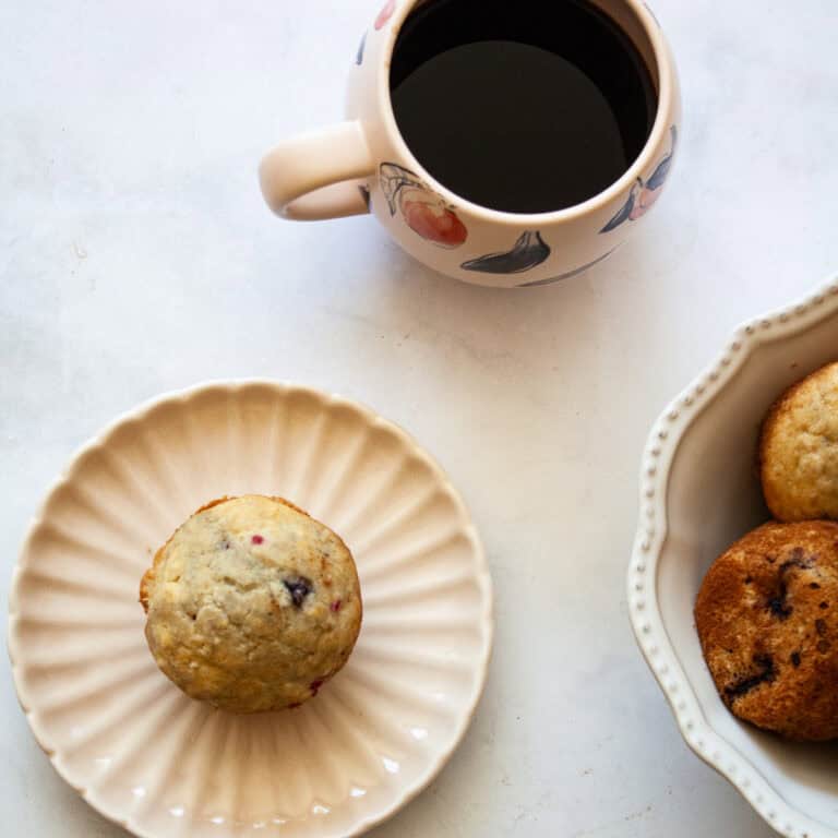 A gluten free raspberry muffin on a plate, a cup of coffee, and a bowl of muffins.