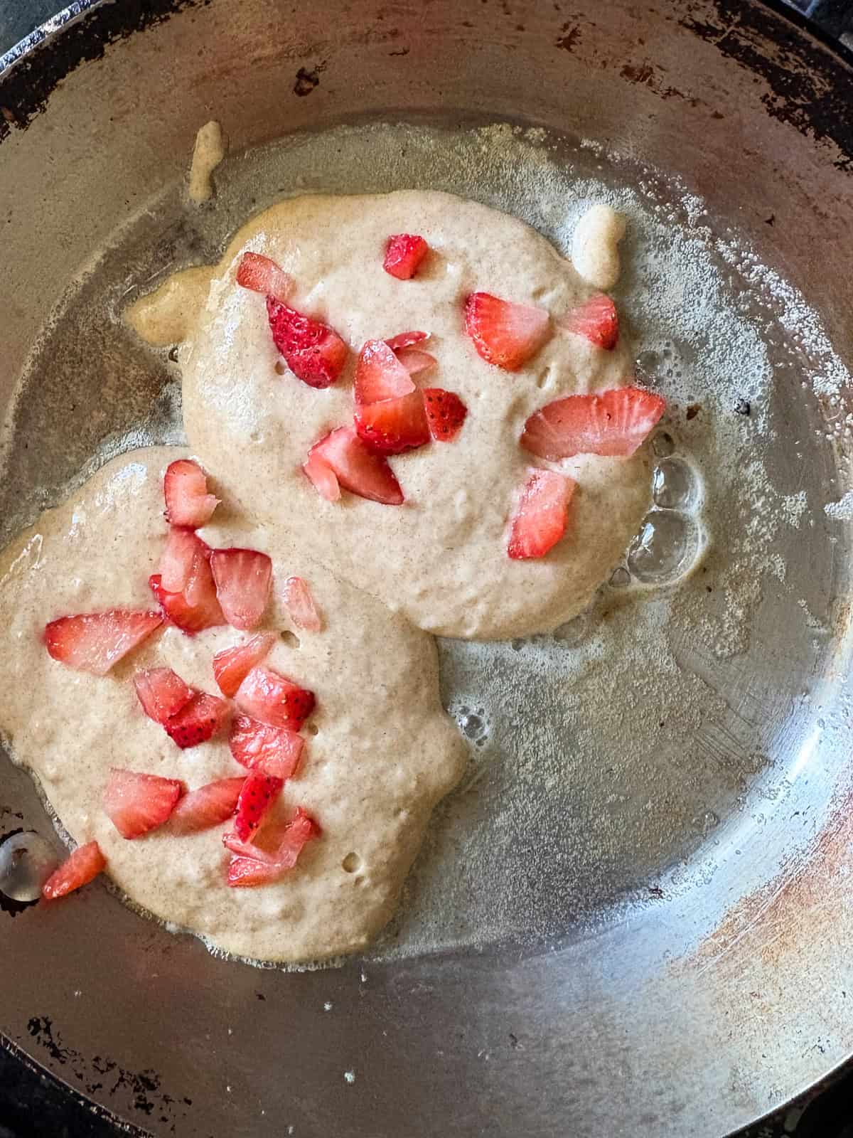 These strawberry pancakes feature fresh, juicy strawberries, whole grain flour, and a light and fluffy crumb. Gluten-free if needed. Two strawberry pancakes cook in a pan.