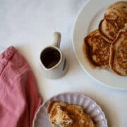 A strawberry pancake and a fork on a plate, with a napkin and a pitcher of maple syrup.
