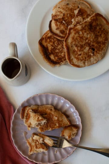 A strawberry pancake and a fork on a plate, with a napkin and a pitcher of maple syrup.