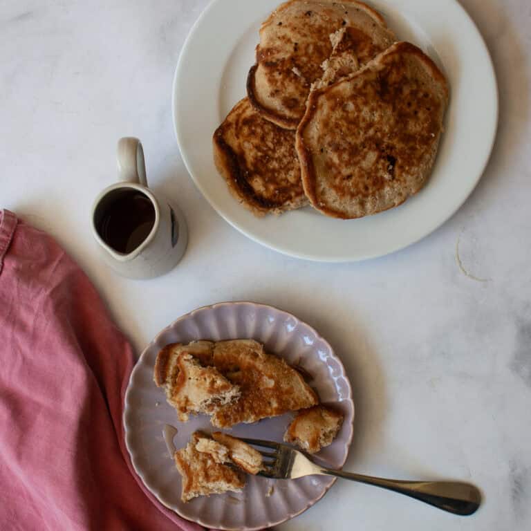 A strawberry pancake and a fork on a plate, with a napkin and a pitcher of maple syrup.