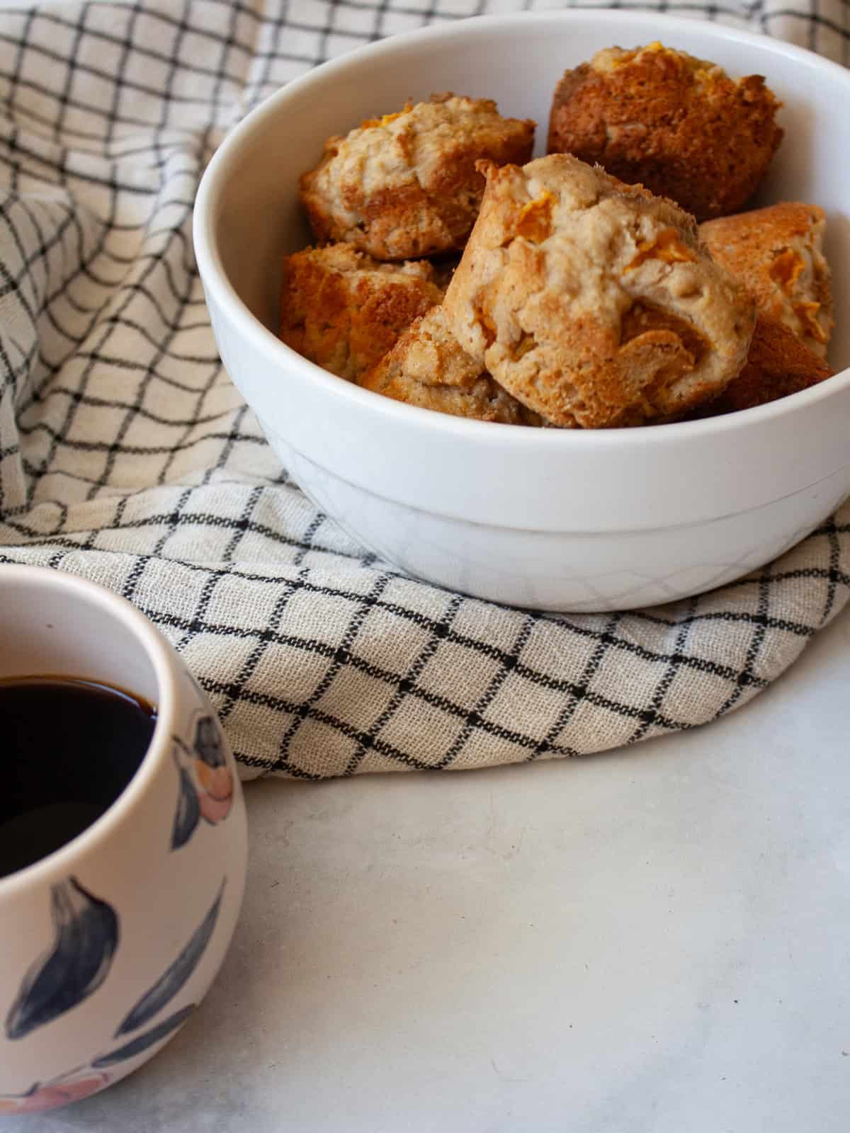 Yogurt, whole grains, and juicy peaches are swirled together with brown sugar in these tender and good-for-you gluten free peach muffins. A bowl of gluten free peach muffins on a checked towel.