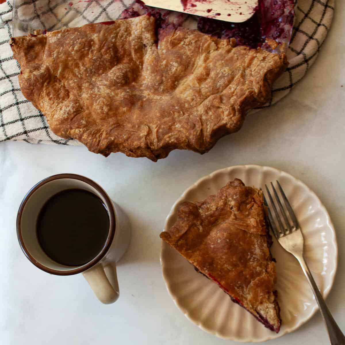A simple method and ingredients makes this blackberry pie an easy, delicious summer bake. Make gluten-free or not, as needed! A slice of blackberry pie on a plate with a fork, with the pie and a cup of coffee next to it.