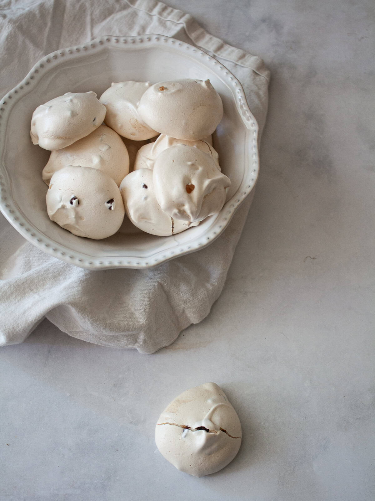 Marshmallowy chocolate chip meringue cookies are studded with mini chocolate chips for bursts of semisweet chocolate in every bite. A bowl of chocolate chip meringue cookies and one in front of it.