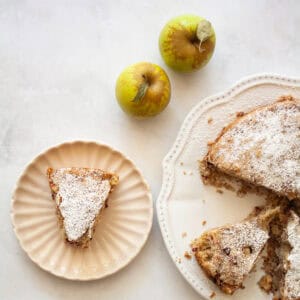 A slice of blackberry apple cake on a plate with apples and the cake next to it.