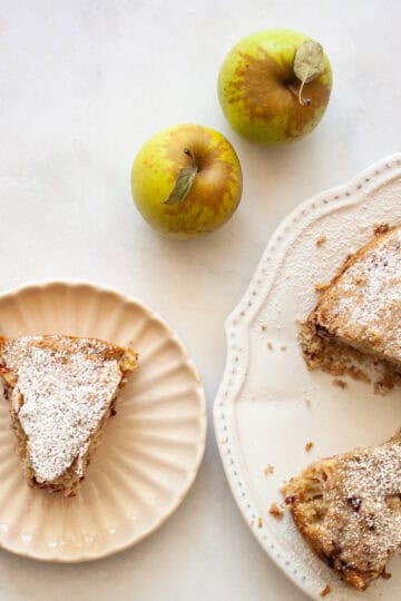 A slice of blackberry apple cake on a plate with apples and the cake next to it.