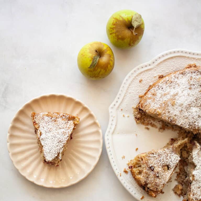 A slice of blackberry apple cake on a plate with apples and the cake next to it.