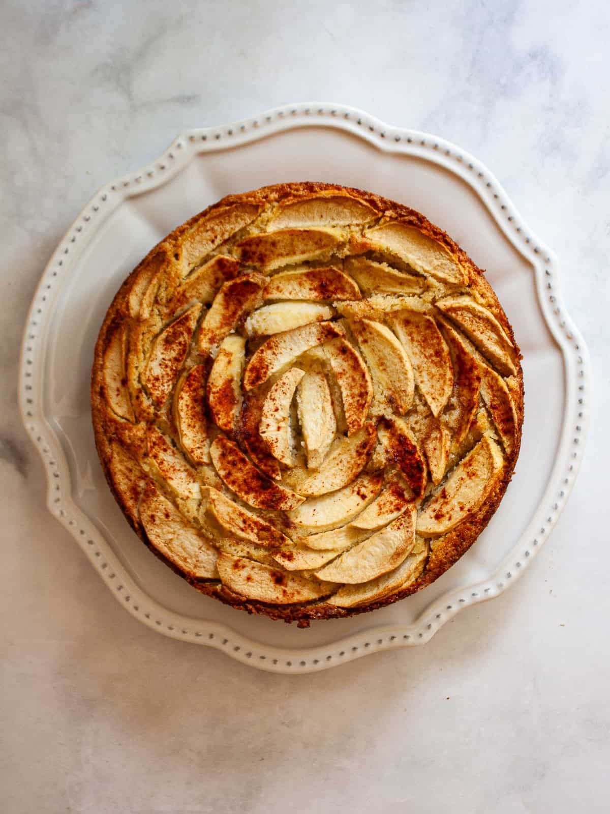 This rich-tasting almond flour apple cake has a wonderfully moist and tender crumb that's topped with sliced apples and maple syrup. An almond flour apple cake on a plate.