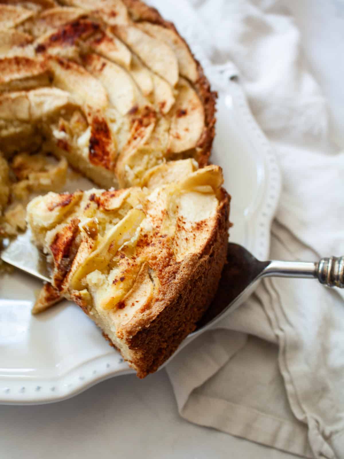This rich-tasting almond flour apple cake has a wonderfully moist and tender crumb that's topped with sliced apples and maple syrup. A server lifts out a slice of almond flour apple cake.