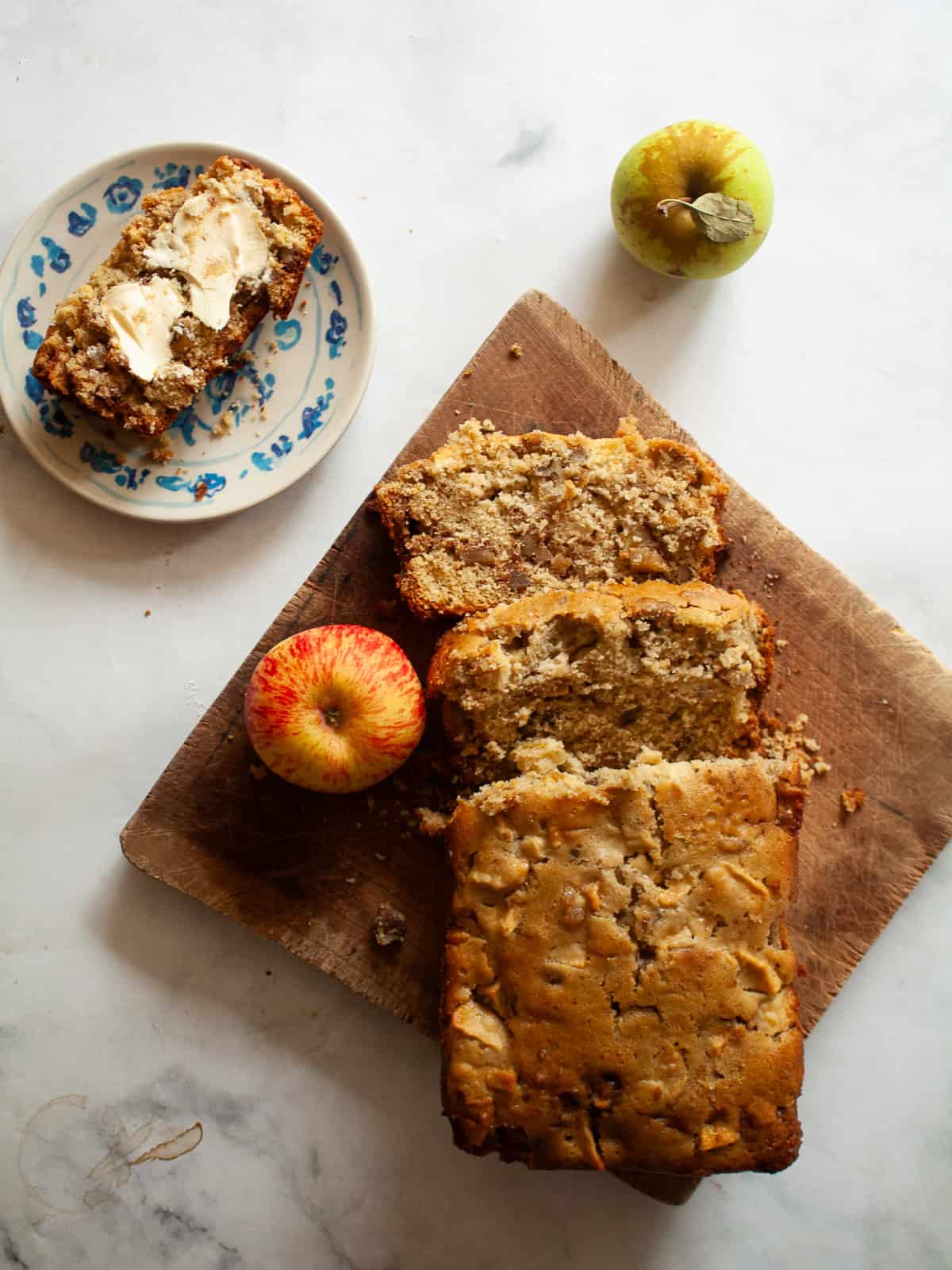 A loaf of apple walnut bread on a cutting board with apples and a slice of bread on a plate.