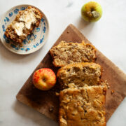 A loaf of apple walnut bread on a cutting board with apples and a slice of bread on a plate.