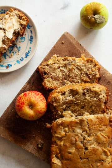 A loaf of apple walnut bread on a cutting board with apples and a slice of bread on a plate.