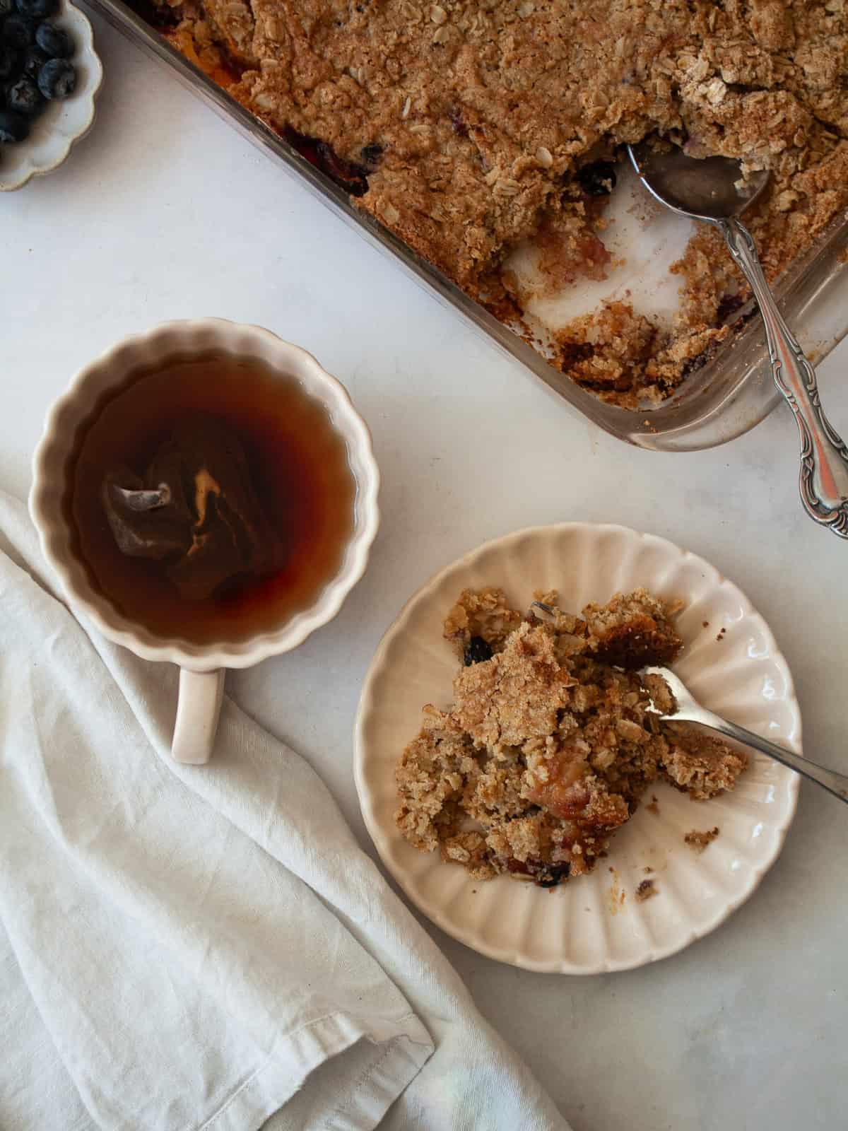 This apple blueberry crisp combines juicy summer apples and berries under a buttery oat topping. A seasonal dessert that's delicious warm. A cup of tea and a portion of apple blueberry crisp and a pan of crisp.
