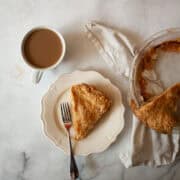 A slice of gluten free apple pie on a plate next to the pie and a cup of coffee.