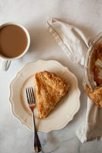 A slice of gluten free apple pie on a plate next to the pie and a cup of coffee.