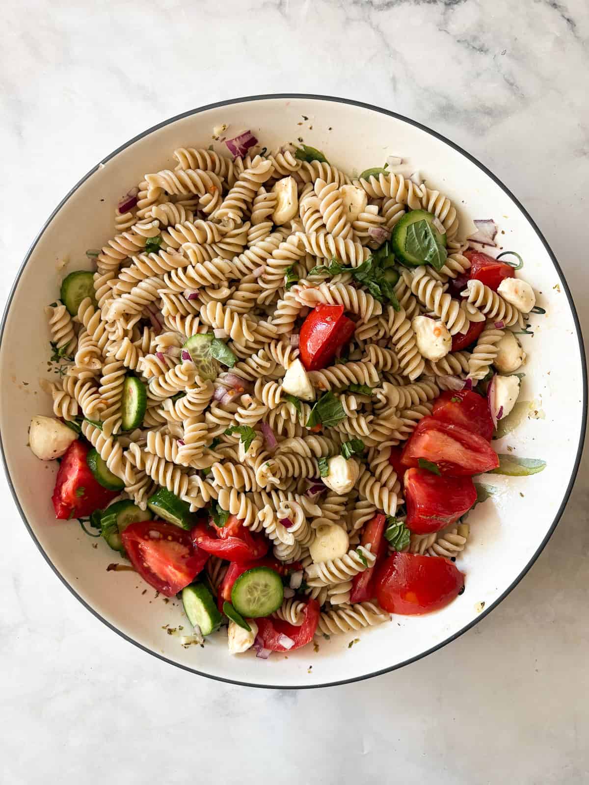 A summer staple, my gluten free pasta salad recipe features a zingy lemon dressing, fresh mozzarella, tomatoes, and fresh herbs. Folding in the herbs to gluten free pasta salad.