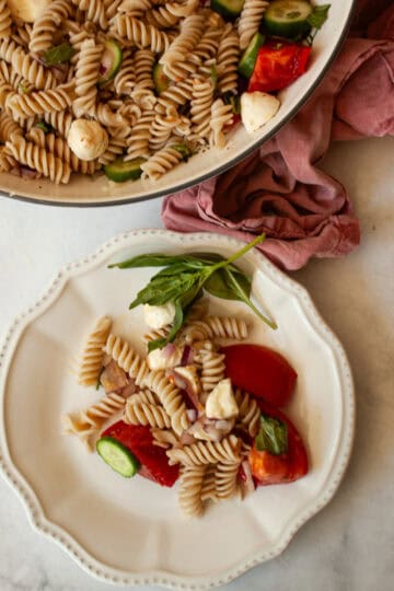 A serving of gluten free pasta salad on a plate and the bowl of salad is seen behind it.