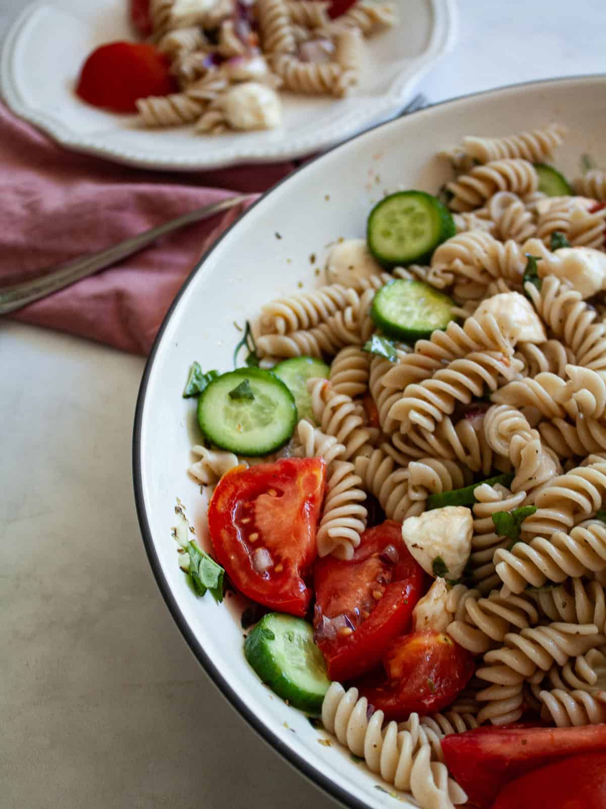 A summer staple, my gluten free pasta salad recipe features a zingy lemon dressing, fresh mozzarella, tomatoes, and fresh herbs. A bowl of gluten free pasta salad shows cucumber and tomatoes.