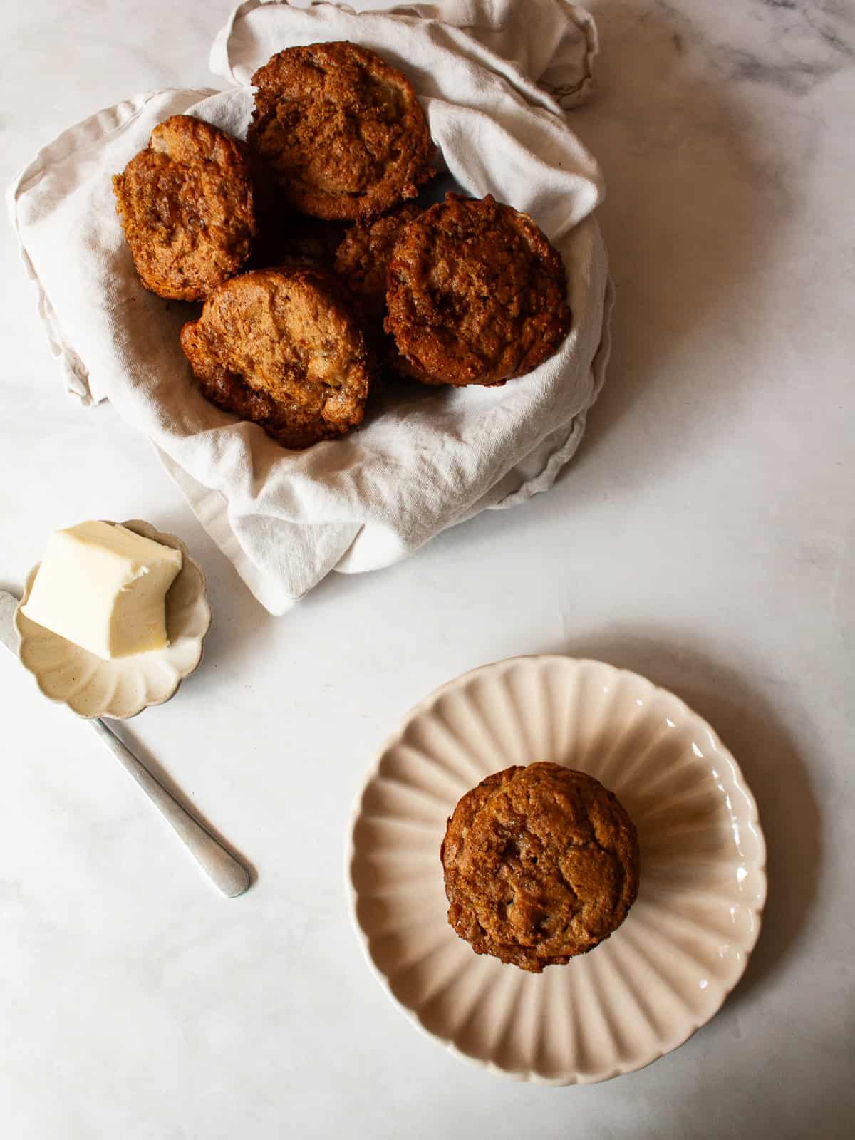 Loaded with fresh, juicy pears, these whole grain pear muffins are studded with walnuts and finished with a crunchy cap of brown sugar. A dish of butter, a pear muffin cut in half on a plate, and a bowl of pear muffins in a napkin.