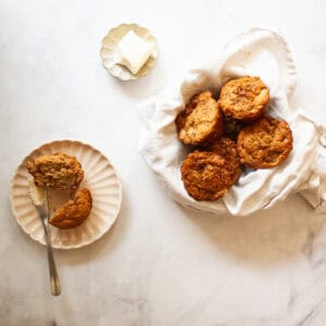 A dish of butter, a pear muffin cut in half on a plate, and a bowl of pear muffins in a napkin.