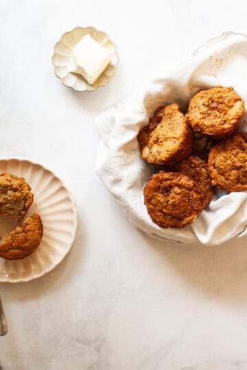 A dish of butter, a pear muffin cut in half on a plate, and a bowl of pear muffins in a napkin.