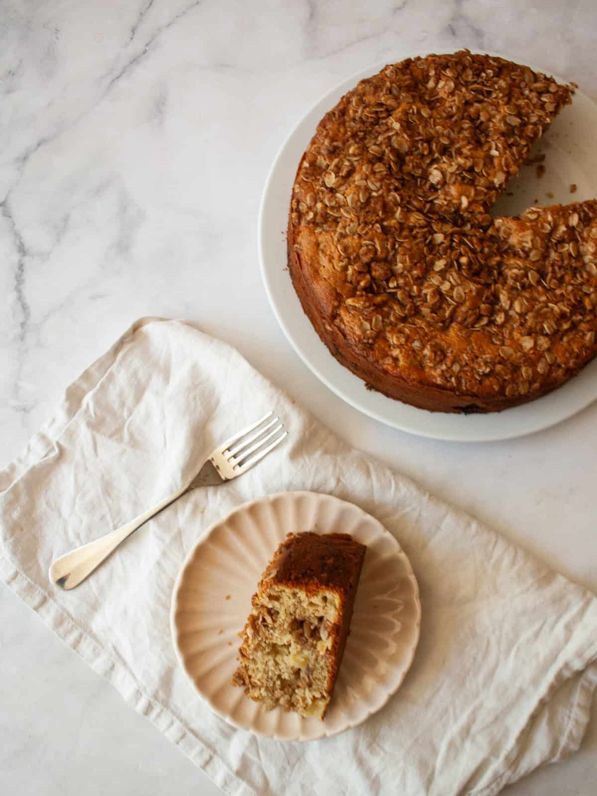 This delicious apple crumb cake is layered with apples, cinnamon, oats, and walnuts. Make it with whole wheat or oat flour for a GF version. An apple crumb cake and a slice of cake on a plate on a napkin with a fork.